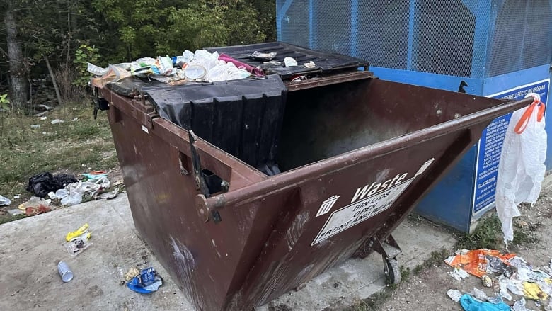 A brown metal garbage bin is overflowing with trash. The lid of the bin is open and damaged. 