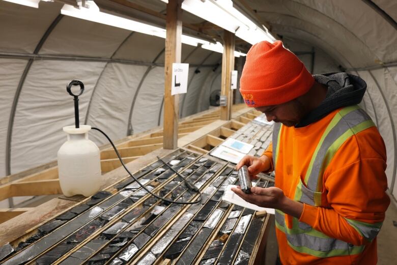 Man in neon construction sweater holds a mineral sample up from a table full of different cut samples in a white tent