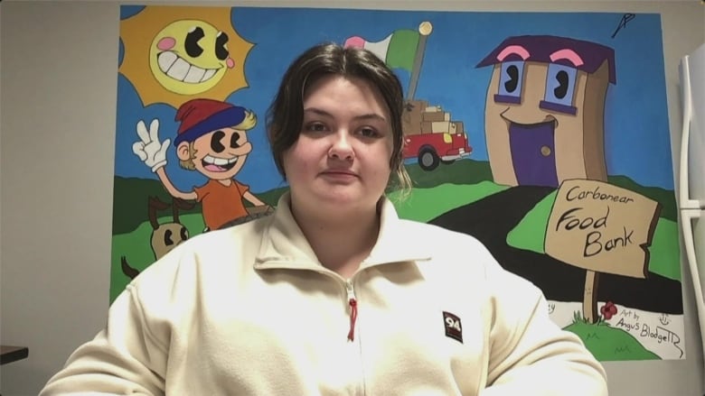 Woman with brown hair pulled back, wearing a white sweater. She's seated and a mural behind her shows a sign that says 