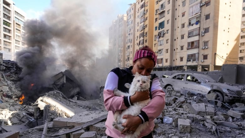 A woman wearing a wrap on her head clutches a white cat while standing amid concrete debris and near a damaged vehicle in a city.