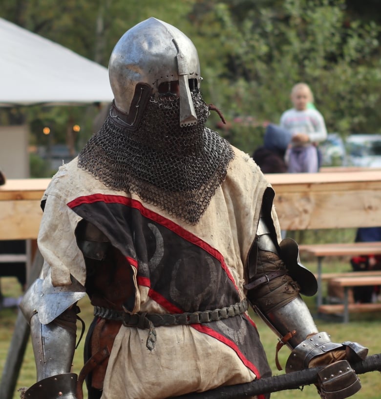 a man in full medieval armour stands holding a battle axe