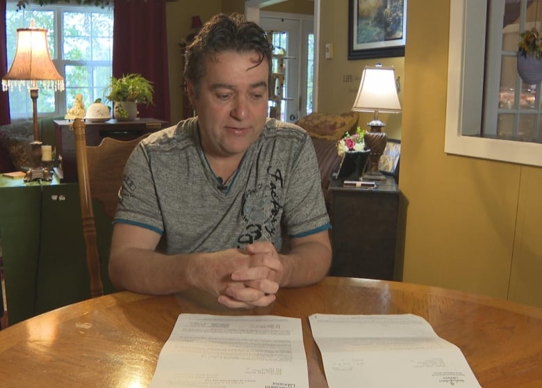 A man wearing a grey tee-shirt sits at his kitchen table. He's looking at two paper documents. 