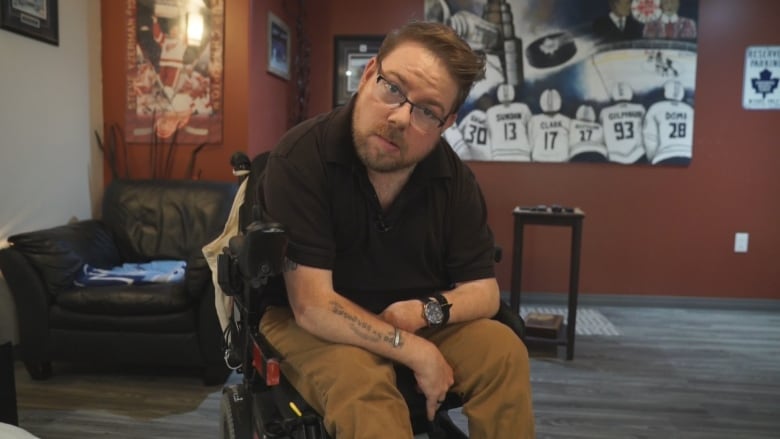 A man with short brown hair and glasses sits in a wheelchair and looks into the camera.