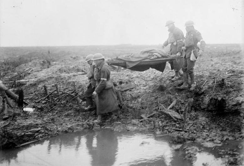 Black and white photo from the First World War shows a wounded soldier being carried across a muddy battlefield.