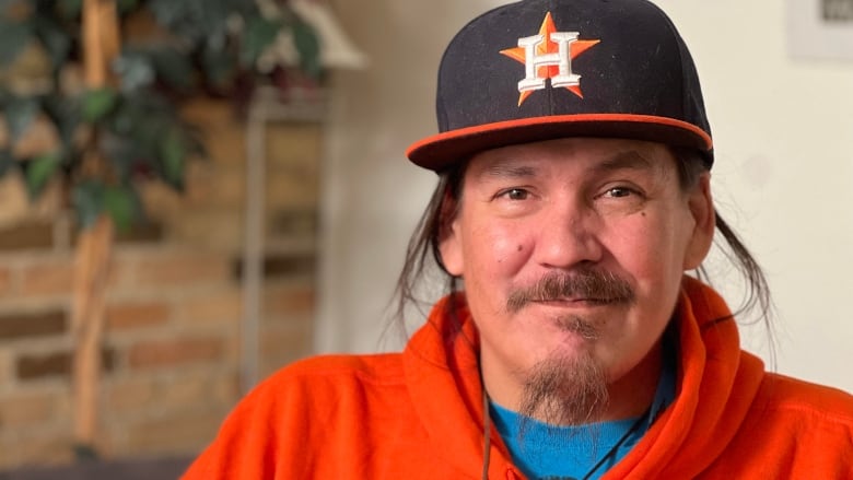 Man with brown hair sits wearing a black and orange baseball hat and orange sweatshirt. 