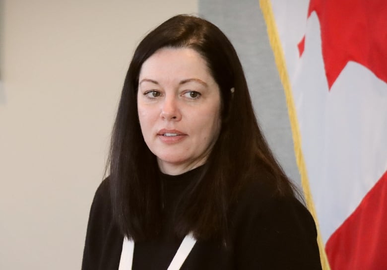 A woman with long black hair wearing a black sweater looks away with a Canadian flag on the wall behind her.