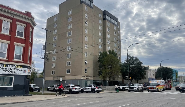 Several police cruisers, an ambulance and a fire truck sit outside a tall apartment building on a city street.