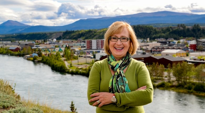 A woman stands in front of a view of Whitehorse, smiling. She is wearing a bright green sweater and wears glasses. 