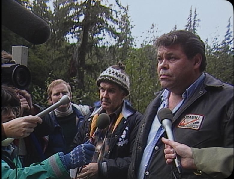 Two men answer questions from a series of media people holding mics and cameras.