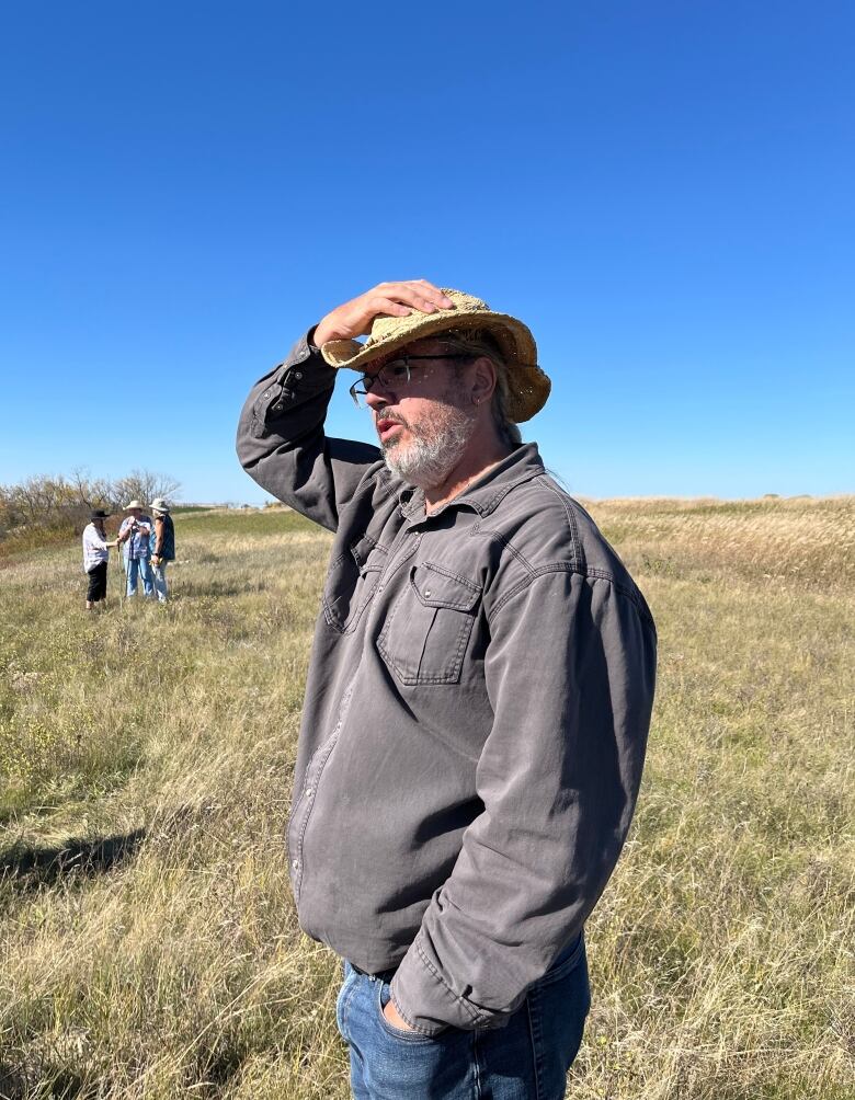 A man in a grey long sleeve shirt stands facing the wind. He hold his straw hat on his head.