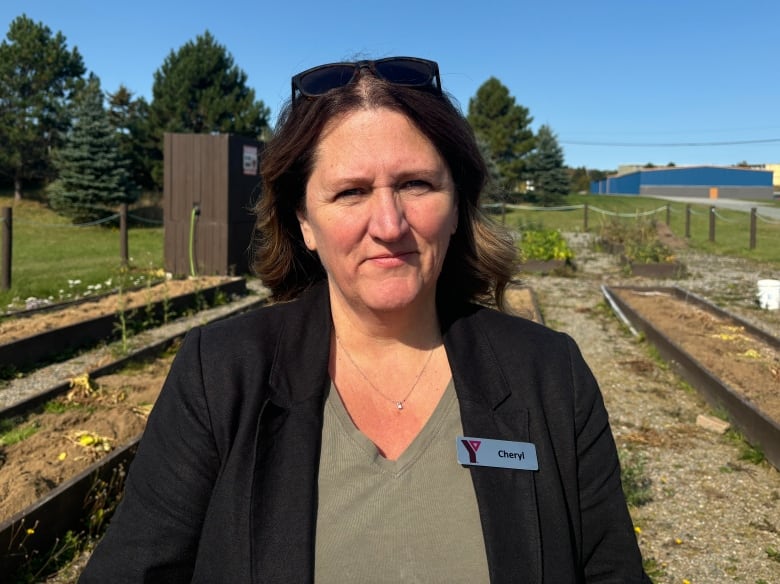 A woman wearing a green shirt and black sweater stands in a garden. She's wearing a nametag that reads 'Cheryl.'