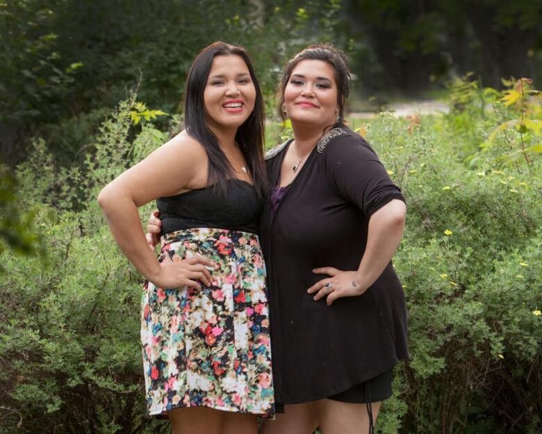 Two women standing together who seem happy with greenery in the back. 