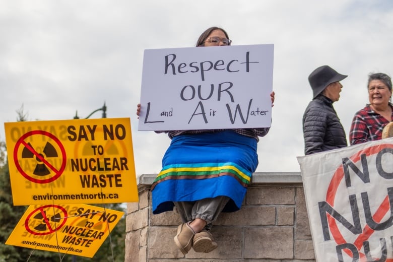 A person is seen sitting on a ledge outside, holding a sign that says 'Respect Our Law: Land, Air, Water.