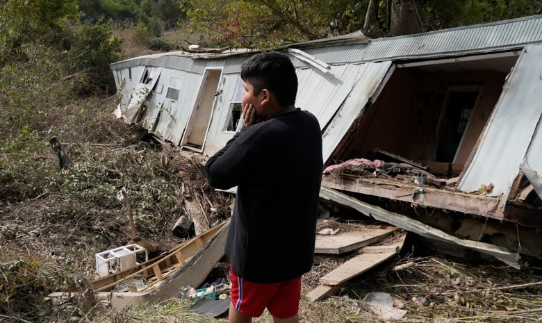 A person stands in the centre of the image, their hands pressed to their face and back to the camera. A white house is leaning on its side, collapsed, with rubble strewn around.