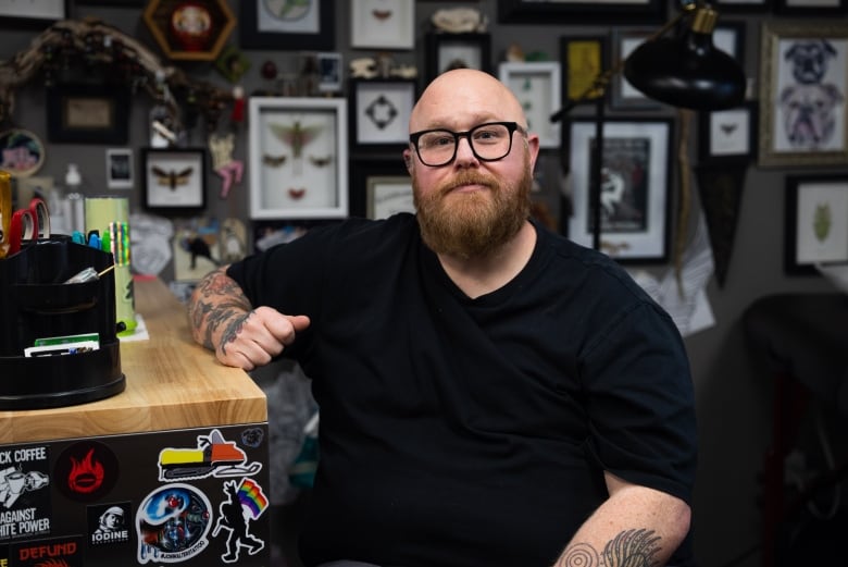 A bearded man with glasses sits inside a tattoo shop.