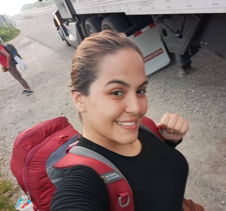 A smiling woman wearing a red backpack takes a selfie on the side of a road next to a large semi truck. 