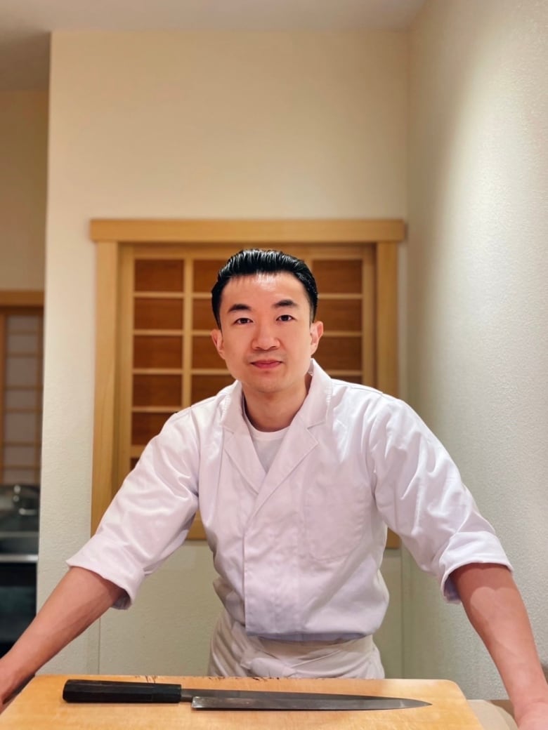 A man leaning against a counter with his hands wearing a white, chef uniform. 