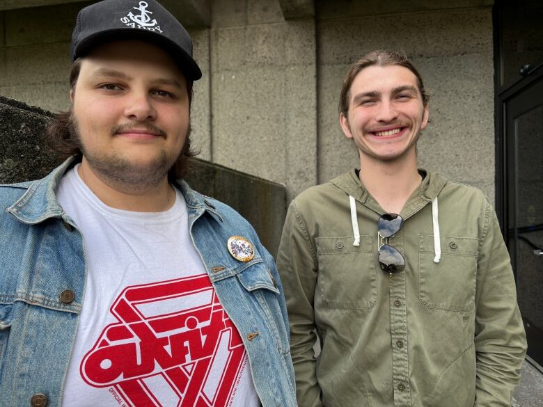 A man with a beard and a baseball cap stands next to a man smiling in a green jacket. They are against a concrete background.