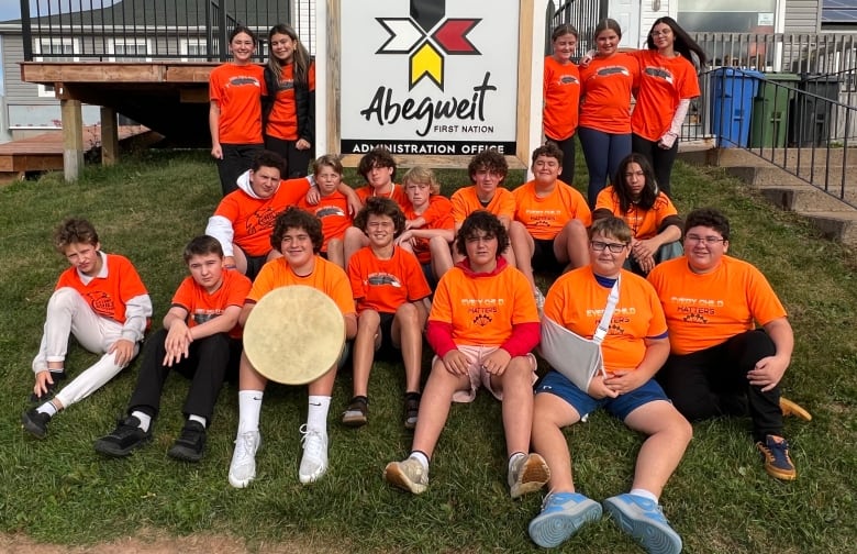 A group of Grade 8 students from Mount Stewart Consolidated School are wearing bright orange shirts. They are sitting in front of the Abegweit First Nation administration office sign.