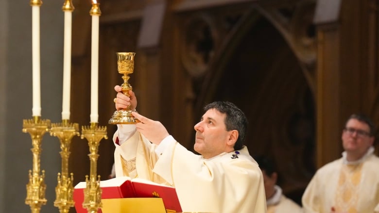 A bishop stands at a pulpit and holds aloft a golden cup. He is white, middle-aged and wearing white robes, looking up to the cup. There are 3 candles in the foreground, and a clergyman behind him.