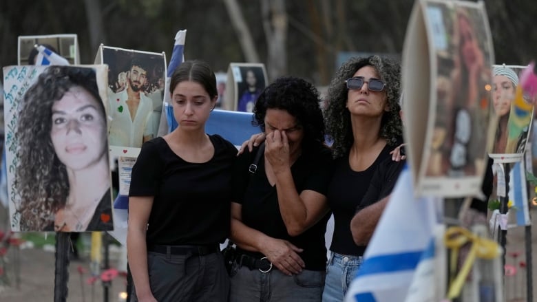 Three women in dark clothing huddle and look mournful, with one appearing to cry, in an outdoor setting that includes Israeli flags and posters depicting other people.