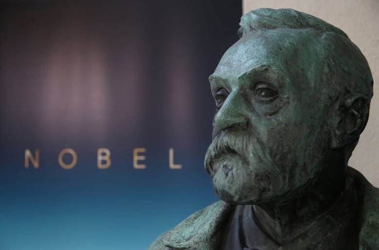 The large, sculpted bust of a bearded man is shown by a wall with an inscription that reads, 'Nobel.'