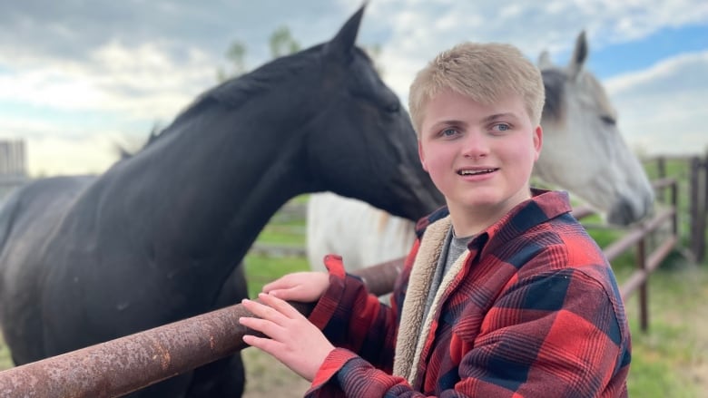 A blonde boy in a red-and-black checked jacket stands next to a black horse.