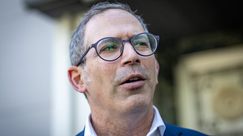 A man with white/brown hair wearing glasses and a blue blazer with a white collared shirt standing in front of a synagogue. 