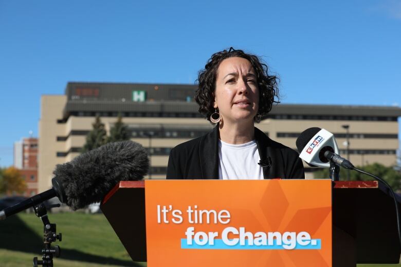 A woman stands behind a podium. The woman is wearing a white shirt and a black jacket. Behind the woman is a hospital. The podium has an orange sign with the phrase 