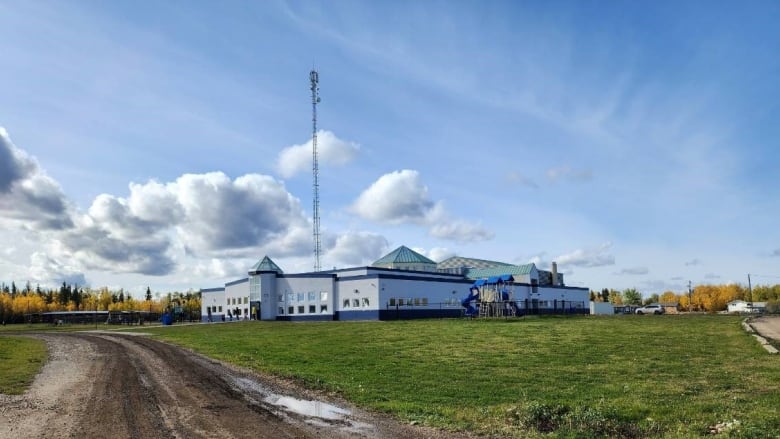 White and blue building in the background, with a dirt road in the foreground. Blue sky with a few white clouds. 