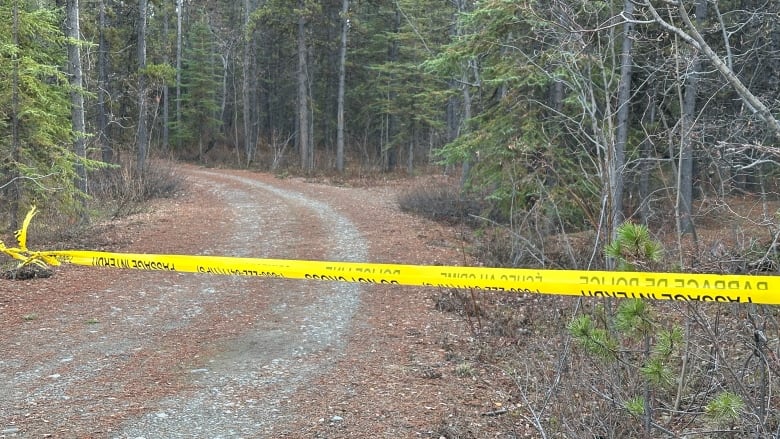 Police tape stretched across a dirt road through a forest.