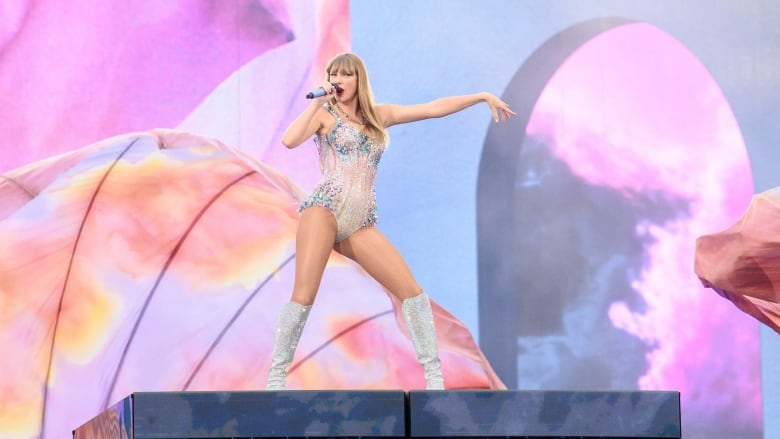 A blonde-haired woman poses with a microphone on a stage against a colourful pastel orange, pink and blue backdrop, wearing a sparkly bodysuit. 