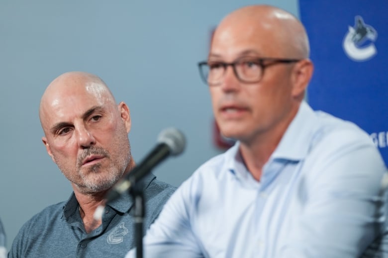 A bald man looks at another bald man who is delivering a news conference