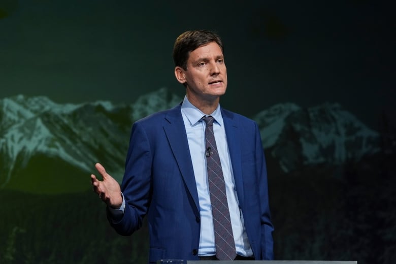 A tall white man points to his right as he speaks in front of a mountain backdrop.