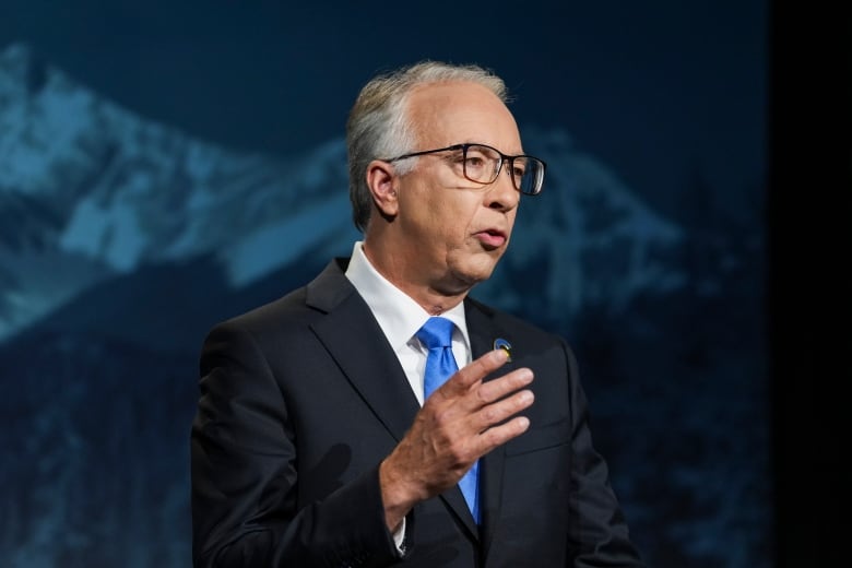 A white man looks to his left as he speaks in front of a mountain backdrop.