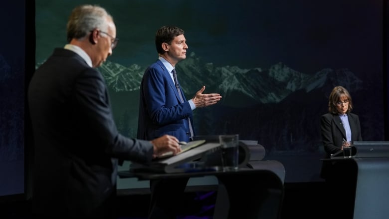 Two men and a woman stand at podiums in front of a mountain backdrop.