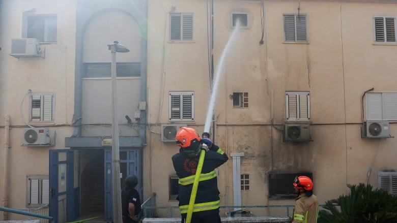 Three firefighters are shown with backs to the camera in full gear and helmets, with one employing a large hose directed at what appears to be a multistorey, low rise building.