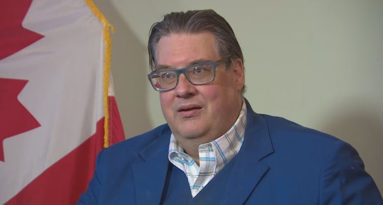 A man in glasses and a blue suit looks to the side of the camera with a Canadian flag behind him.