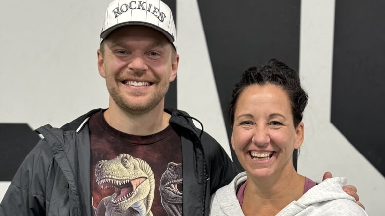 A bearded man in a ball cap and a smiling woman in a hoodie stand with their arms around each other.