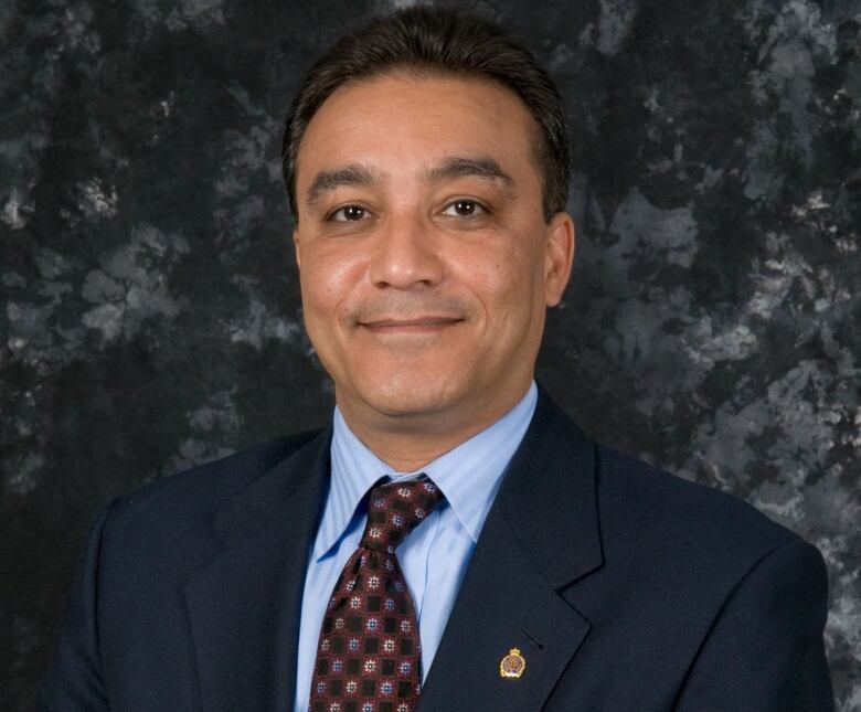 A headshot of a man in a navy suit and red tie, wearing police pins.