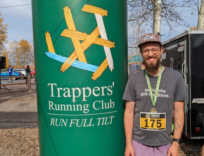 A smiling man wearing a runners bib and medal stands in front of a pole showing the logo for the Trappers' Running Club.