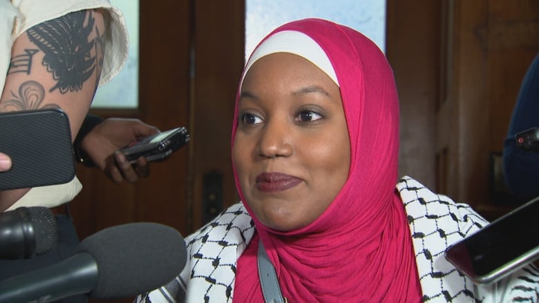 A portrait of a person in a hijab and keffiyeh speaking into a cluster of microphones.