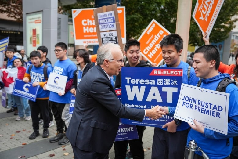 A man shakes hands with young, mostly Asian men waving blue Conservative placards.