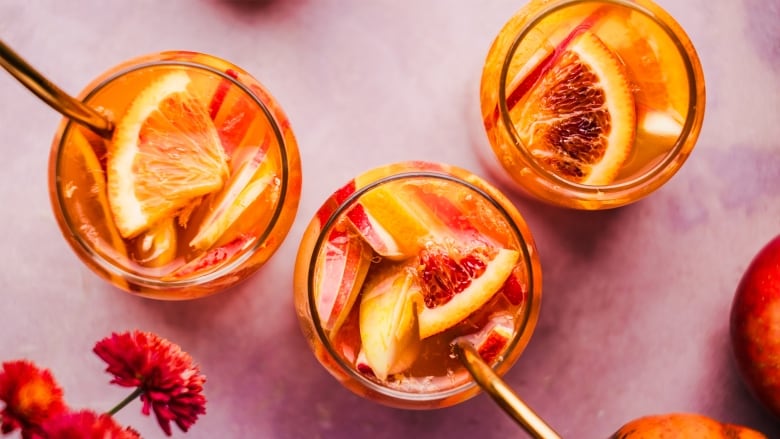 Overhead shot of 3 glasses of an orange cocktail with slices of apple and orange in them, all sitting on a pink background.  