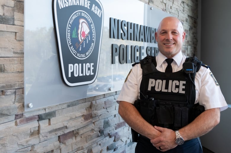 A bald man wearing a white short-sleeved shirt with epaulettes and a black vest with 