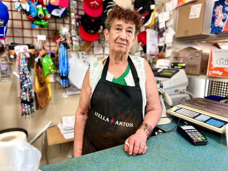 A woman wearing a black apron stands in front of a counter with a cash register on it. 