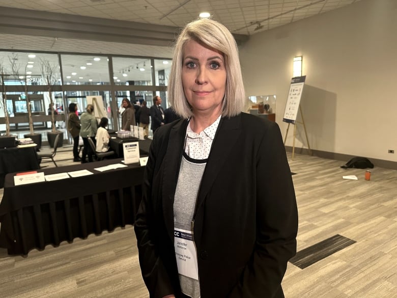 A woman with blond hair and a suit stands in front of a table.