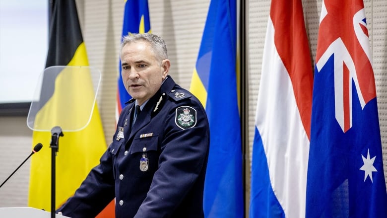 A man with short grey hair, dressed in a police uniform, stands in front of a podium.