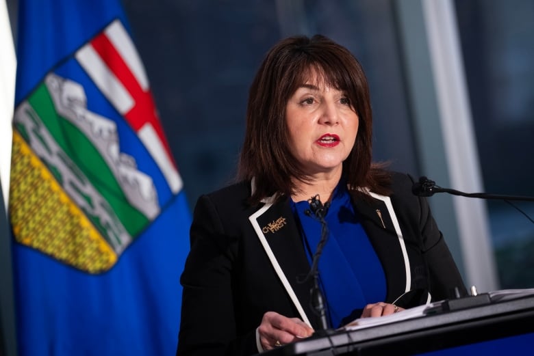 A woman wearing a blazer stands in front of a flag while speaking at a podium.