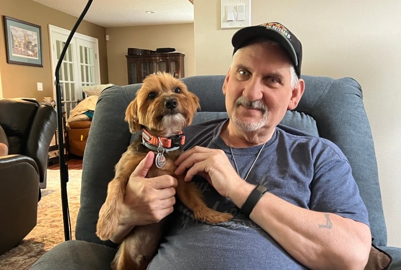 A man in a blue T-shirt and black ball cap holds a dog while he sits in an easy chair.